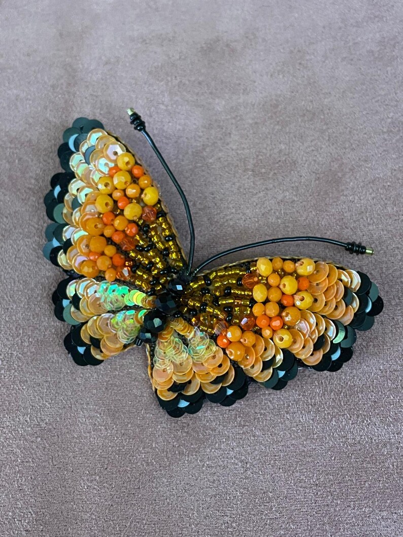 May include: A beaded butterfly brooch with black, orange, and yellow sequins. The butterfly has a black wire attached to the back for pinning.
