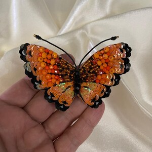 May include: A beaded butterfly brooch with orange, black, and iridescent sequins. The butterfly is detailed with black beading and has a wire pin on the back.