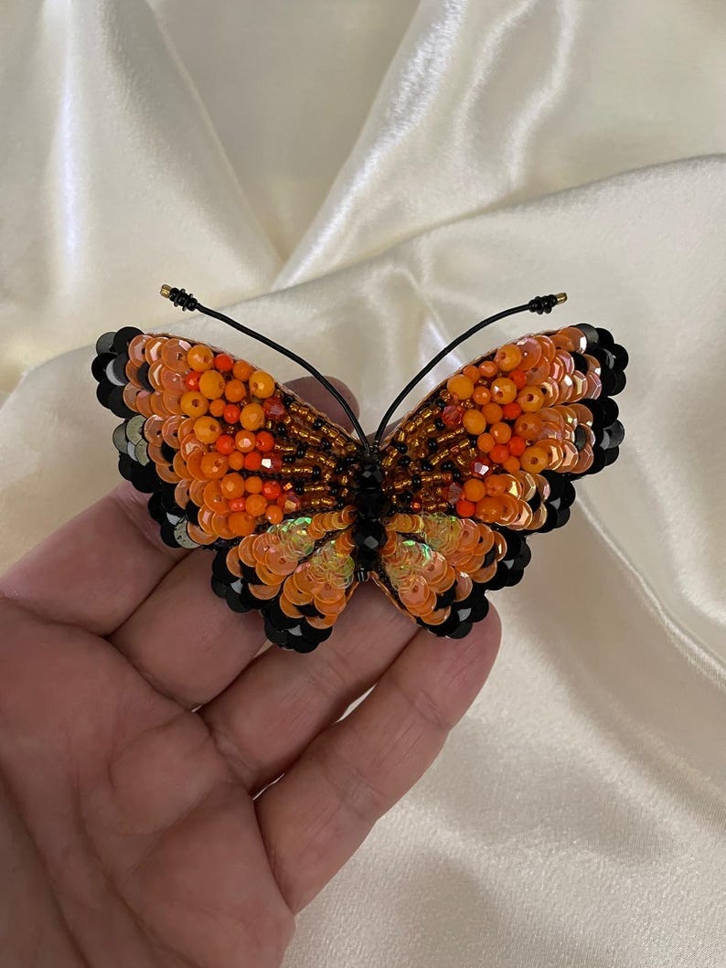 May include: A beaded butterfly brooch with orange, black, and iridescent sequins. The butterfly is detailed with black beading and has a wire pin on the back.