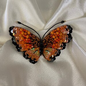 May include: A beaded butterfly brooch with orange, red, and black sequins. The butterfly is made of iridescent sequins and beads.