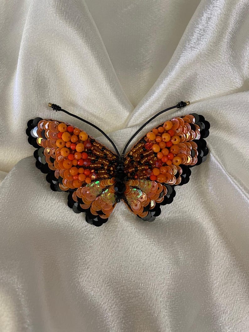 May include: A beaded butterfly brooch with orange, red, and black sequins. The butterfly is made of iridescent sequins and beads.