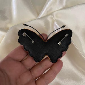 May include: Black felt butterfly brooch with a gold wire and safety pin closure. The butterfly has a scalloped edge and is decorated with orange and yellow embroidery.