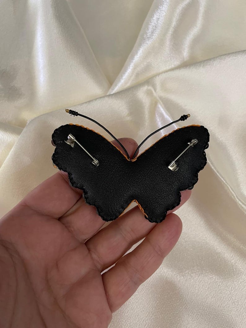May include: Black felt butterfly brooch with a gold wire and safety pin closure. The butterfly has a scalloped edge and is decorated with orange and yellow embroidery.