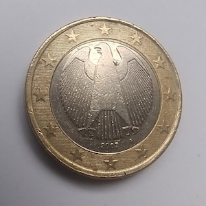 May include: A one euro coin featuring a detailed depiction of a German eagle. The coin has a gold-colored outer ring with stars and a silver-colored center. The year 2002 is visible on the coin.