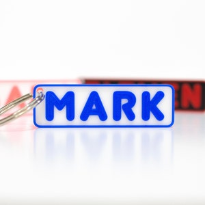 Puede incluir: Un llavero rectangular blanco con la palabra "MARK" en letras azules. El llavero está unido a un anillo plateado.