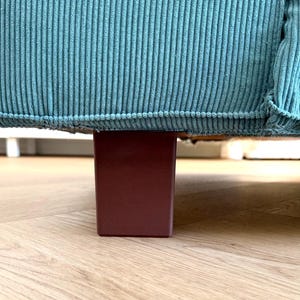 May include: Close-up of a section of a teal corduroy sofa. The sofa sits on a rectangular, burgundy-colored wooden leg. The floor is light-colored wood.