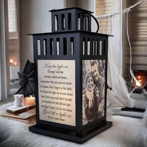 May include: A black metal lantern featuring a square frame with vertical slats. One side displays the text: Keep the light on, Though God has already called me home, remember that you are not alone. Another side features a black and white photograph.