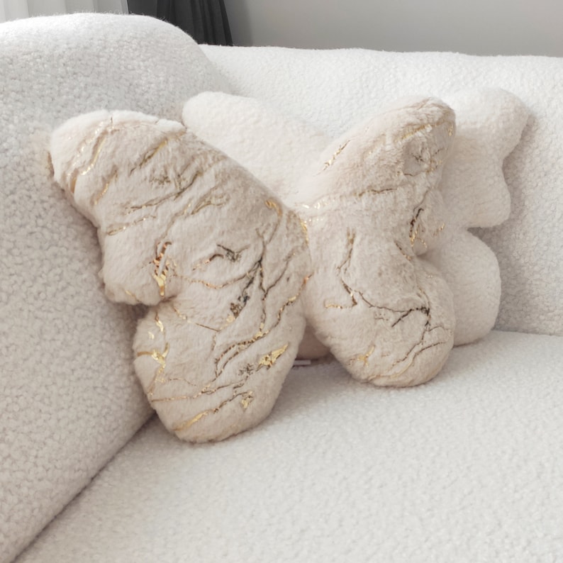 May include: A butterfly-shaped decorative pillow in cream and beige tones. The plush pillow features gold-colored vein-like accents across its surface. The pillow is placed on a white, textured sofa, creating a soft and inviting aesthetic.