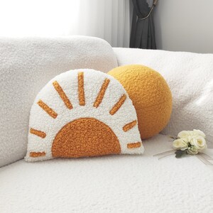May include: A decorative pillow set featuring a sun-shaped pillow in white and orange, and a round yellow pillow. The pillows are made of a soft, textured material and are placed on a white couch. The sun pillow has a half-circle orange center with radiating lines.