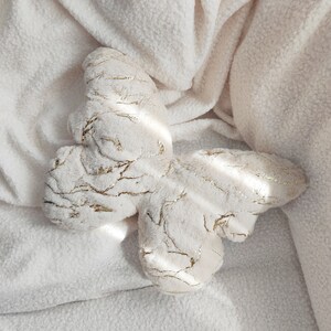 May include: A plush butterfly-shaped pillow in cream with gold metallic accents. The butterfly is lying on a cream-colored, textured blanket. The pillow's design features gold veins throughout the cream fabric.