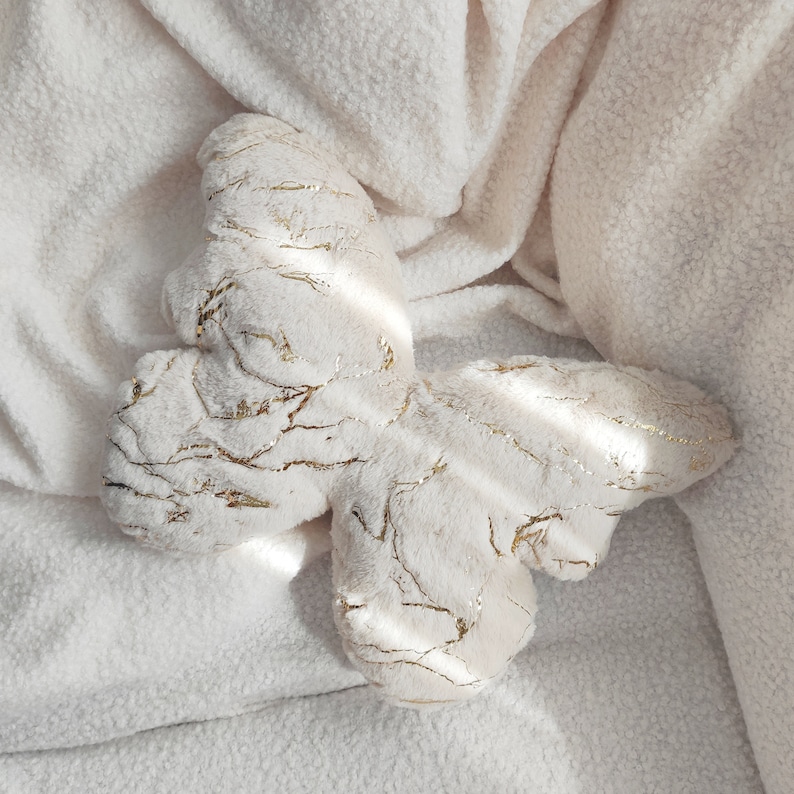 May include: A plush butterfly-shaped pillow in cream with gold metallic accents. The butterfly is lying on a cream-colored, textured blanket. The pillow's design features gold veins throughout the cream fabric.