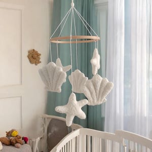 May include: A white nursery mobile with plush seashell and starfish shapes hanging from a wooden ring. The mobile is suspended by white string, creating a calming, ocean-themed decoration for a baby's room.
