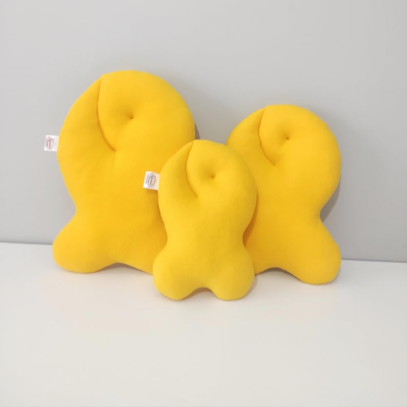 May include: Three bright yellow, plush, abstract-shaped pillows of varying sizes. Each pillow has a rounded top and a unique, three-lobed base. Small white tags are attached to the side of the largest and smallest pillows.