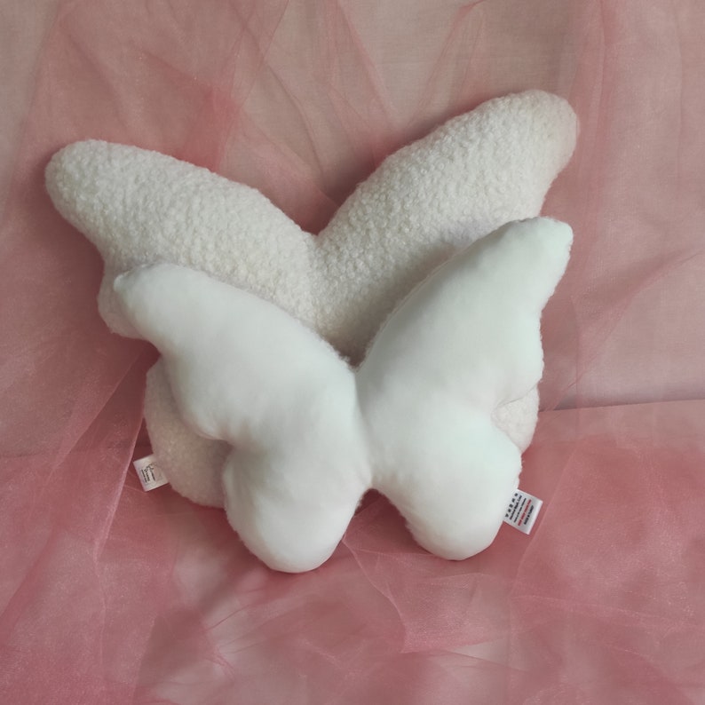 May include: A white butterfly-shaped pillow with two sets of wings. The top set of wings has a textured, fluffy appearance, while the bottom set is smooth. The pillow is on a pink surface.