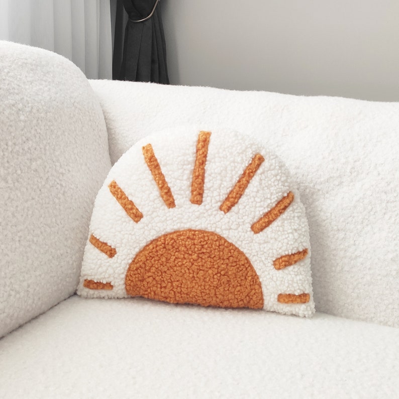 May include: A white, half-circle pillow with an orange sun design. The pillow has a textured, fluffy appearance. The sun's rays and center are a warm orange color, contrasting with the white background. The pillow is placed on a white couch.