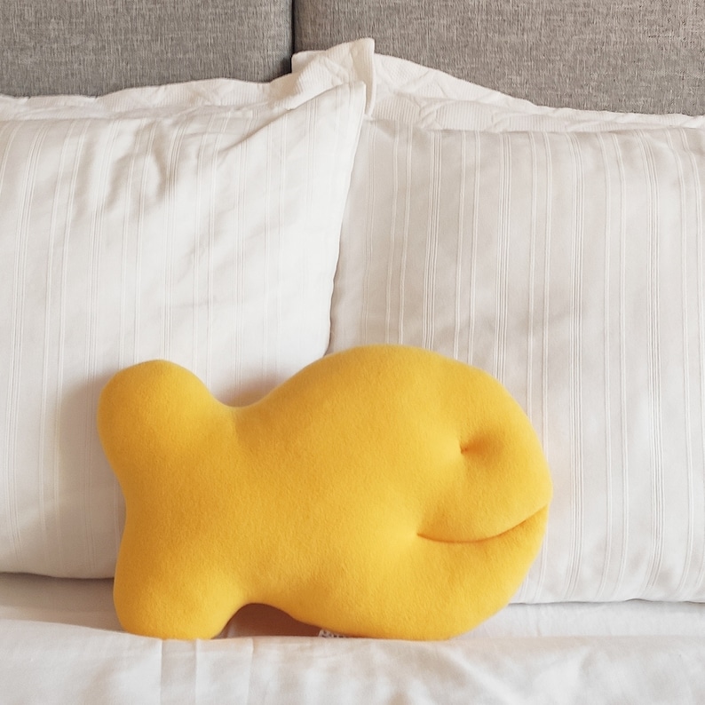 May include: A bright yellow, fish-shaped decorative pillow. The pillow is made of soft fabric and rests on a white bed with two white pillows and a gray headboard in the background.