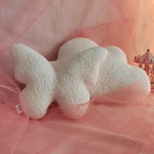 May include: A fluffy, white cloud-shaped pillow made of soft, textured fabric. The pillow has a three-dimensional design with rounded edges and a plush appearance. The background is a soft pink fabric, creating a dreamy aesthetic.