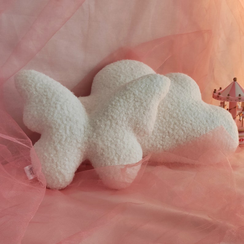 May include: A fluffy, white cloud-shaped pillow made of soft, textured fabric. The pillow has a three-dimensional design with rounded edges and a plush appearance. The background is a soft pink fabric, creating a dreamy aesthetic.