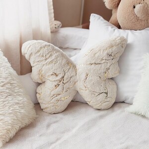 May include: A cream-colored butterfly-shaped pillow with gold accents. The plush pillow is resting on a white bed with other pillows and a teddy bear in the background. The butterfly pillow has a textured surface.