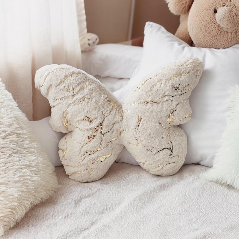 May include: A cream-colored butterfly-shaped pillow with gold accents. The plush pillow is resting on a white bed with other pillows and a teddy bear in the background. The butterfly pillow has a textured surface.