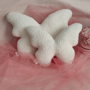 May include: Three white butterfly-shaped pillows with a textured, plush surface. The pillows are arranged on a pink tulle fabric, with a miniature pink carousel in the background. The pillows are a soft, decorative item.