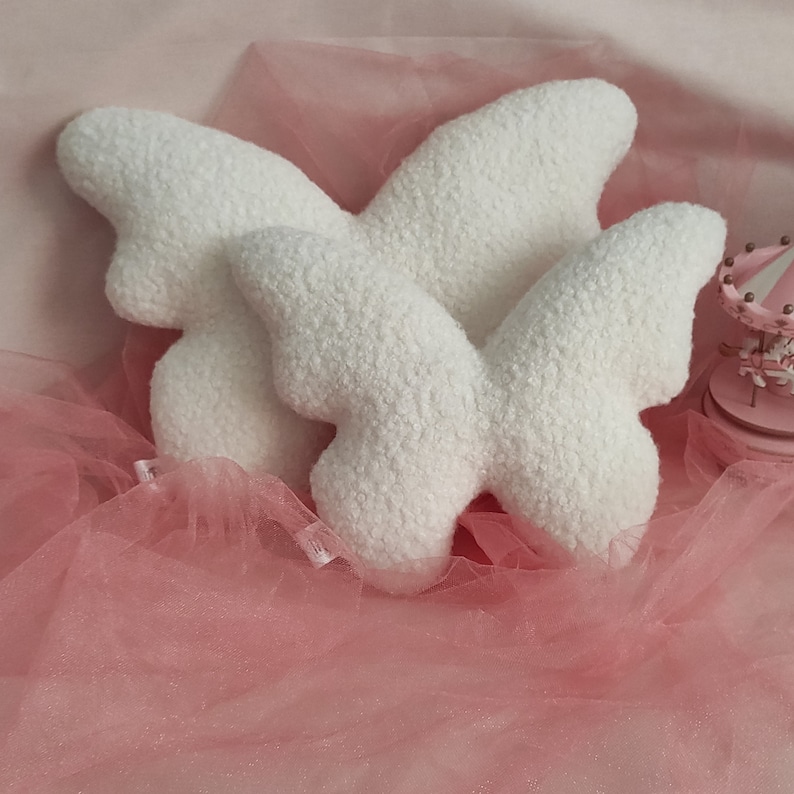May include: Three white butterfly-shaped pillows with a textured, plush surface. The pillows are arranged on a pink tulle fabric, with a miniature pink carousel in the background. The pillows are a soft, decorative item.