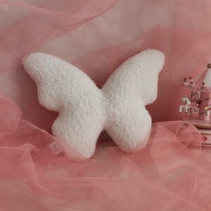 May include: A white, plush butterfly-shaped pillow with a textured surface. The pillow is set against a pink backdrop with a carousel in the background. The butterfly pillow is a decorative item.