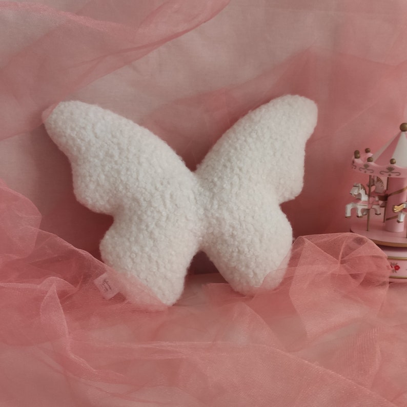 May include: A white, plush butterfly-shaped pillow with a textured surface. The pillow is set against a pink backdrop with a carousel in the background. The butterfly pillow is a decorative item.