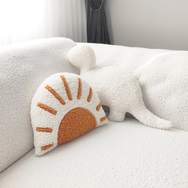 May include: A white, textured sofa with two decorative pillows. One pillow is shaped like a sun with an orange center and rays. The other pillow is shaped like a dinosaur. The pillows are made of a soft, plush material.