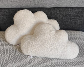 May include: Two fluffy, white cloud-shaped decorative pillows. The pillows have a soft, textured surface and are placed on a gray patterned surface. The pillows are a light cream color and are designed to look like clouds.