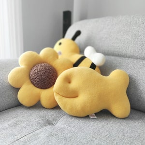 May include: A collection of yellow plush pillows in the shapes of a sunflower, a bee, and a fish. The sunflower pillow has a brown center. The bee pillow has black stripes and white wings. The fish pillow has a smiling face.