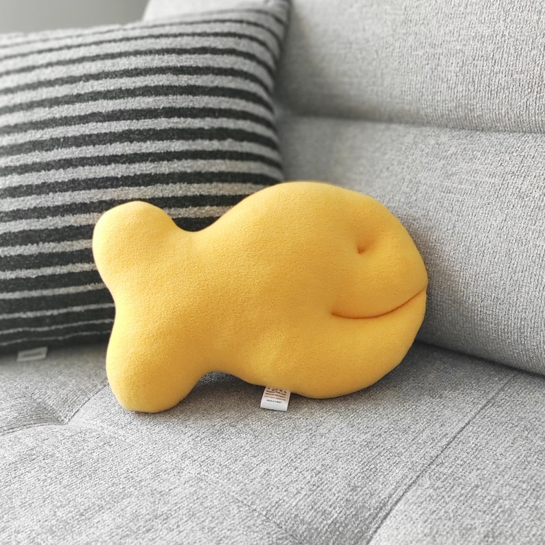 May include: A bright yellow, fish-shaped decorative pillow. The pillow has a smiling face and is made of a soft, textured material. It is resting on a gray couch next to a striped pillow.