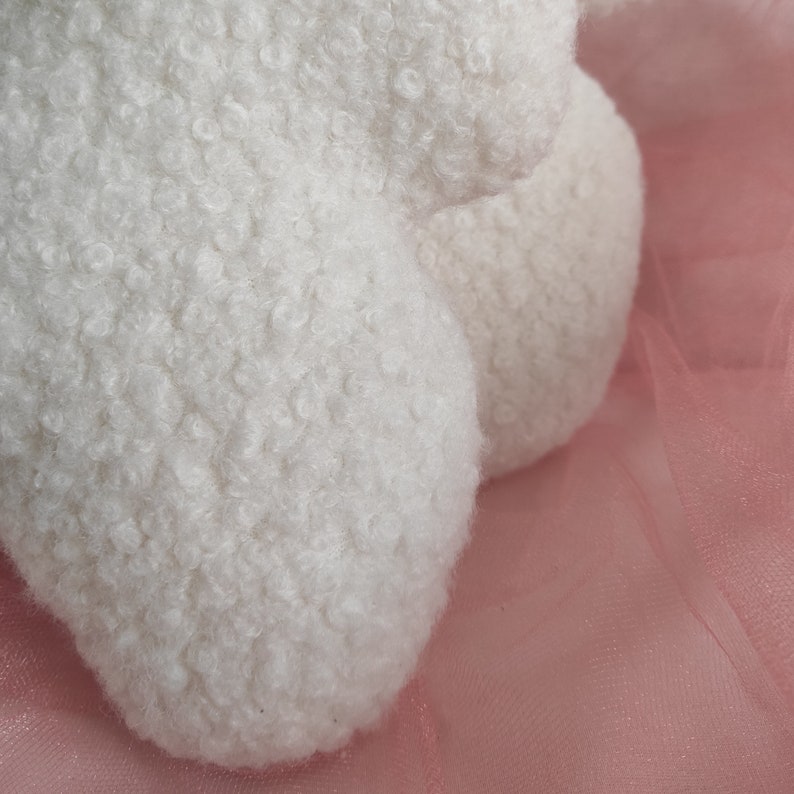May include: Close-up of a white, fluffy, textured object, possibly a stuffed animal or decorative item. The material has a dense, looped pile, creating a soft, cloud-like appearance. The background is a soft pink, possibly tulle fabric.