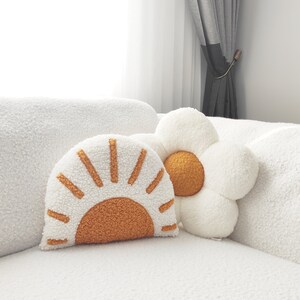 May include: Two decorative throw pillows. One is shaped like a sun with a white background and orange rays. The other is a white flower with an orange center. Both pillows have a soft, textured appearance and are placed on a white couch.