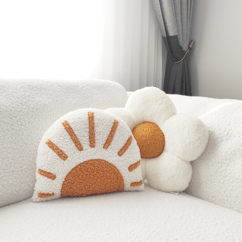 May include: Two decorative throw pillows. One is shaped like a sun with a white background and orange rays. The other is a white flower with an orange center. Both pillows have a soft, textured appearance and are placed on a white couch.
