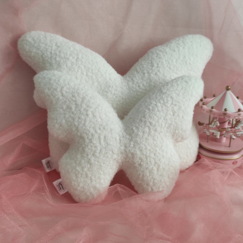 May include: A pair of fluffy white butterfly-shaped pillows. The pillows have a soft, textured appearance and are made of a plush material. A small pink carousel is in the background, adding a whimsical touch to the scene.