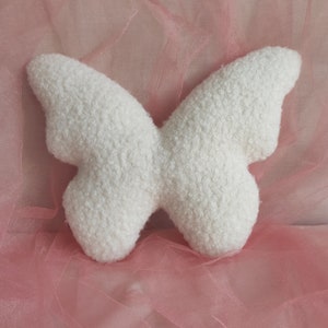 May include: A white, butterfly-shaped pillow made of a textured, fluffy material. The pillow is set against a soft pink background, creating a delicate and whimsical aesthetic. The butterfly design is symmetrical and detailed.