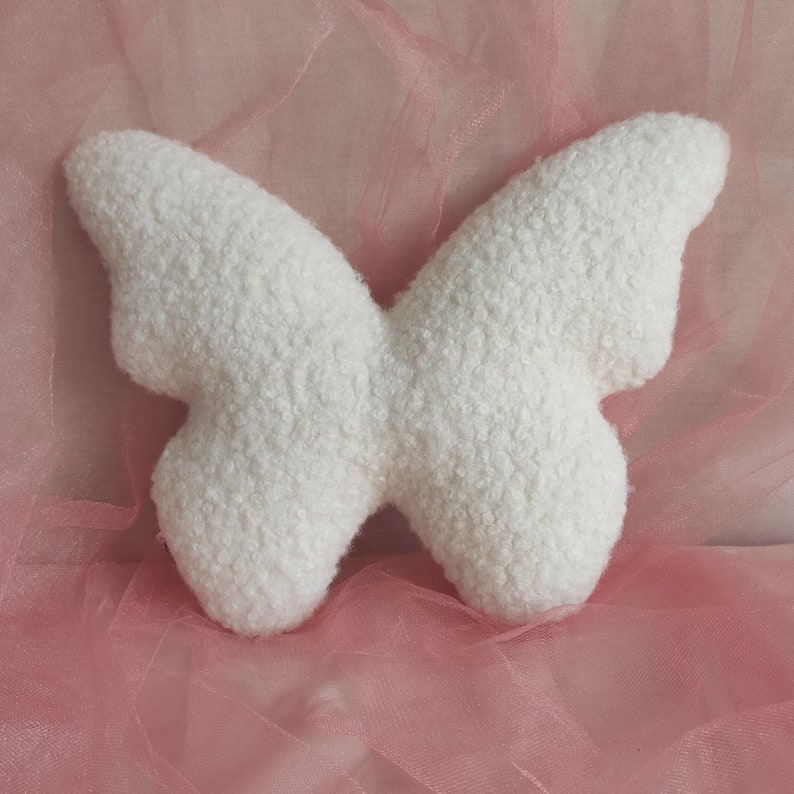 May include: A white, butterfly-shaped pillow made of a textured, fluffy material. The pillow is set against a soft pink background, creating a delicate and whimsical aesthetic. The butterfly design is symmetrical and detailed.