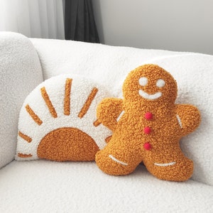 May include: Two decorative throw pillows on a white couch. One pillow is shaped like a gingerbread man with red buttons and white accents. The other pillow is a half-circle with a sun design in white and orange.