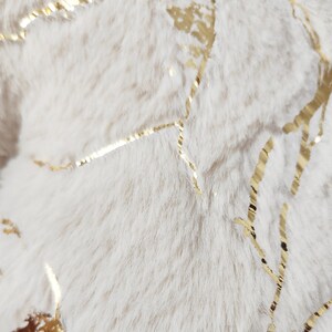 May include: Close-up of a soft, off-white faux fur fabric with gold metallic accents. The gold accents are in a vein-like pattern, creating a luxurious and textured appearance. The fur has a plush, fluffy texture.
