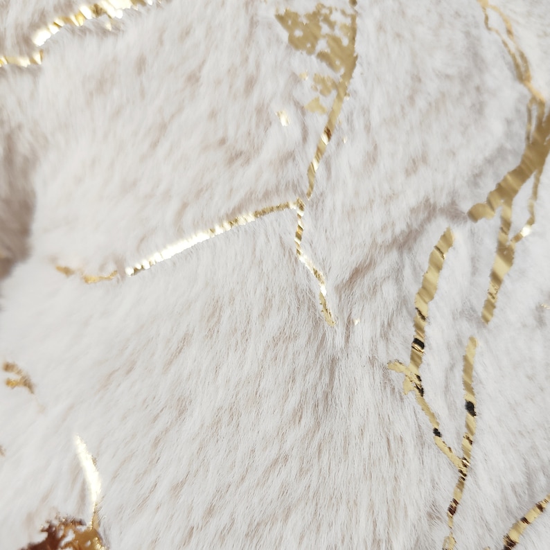 May include: Close-up of a soft, off-white faux fur fabric with gold metallic accents. The gold accents are in a vein-like pattern, creating a luxurious and textured appearance. The fur has a plush, fluffy texture.