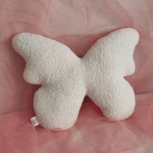 May include: A white butterfly-shaped pillow with a textured, plush surface. The pillow is made of a soft material and is designed to resemble a butterfly. The wings are symmetrical and rounded, creating a whimsical decorative item.