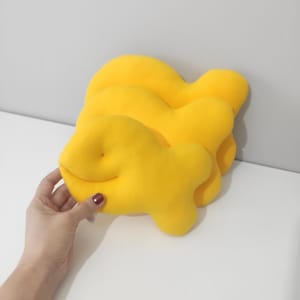 May include: Three bright yellow, plush fish-shaped pillows. One pillow is being held by a hand. The pillows have a soft, rounded shape and a smiling face. The background is a plain white surface.