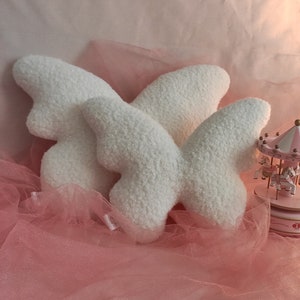 May include: Three fluffy, white butterfly-shaped pillows rest on a pink tulle fabric. The pillows have a soft, textured appearance. A small, pink carousel is visible in the background, adding a whimsical touch to the scene.