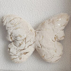 May include: A plush butterfly-shaped pillow in cream with gold metallic accents. The pillow has a soft, textured surface and a detailed design. The wings are spread out, and the gold accents create a striking contrast against the cream color.