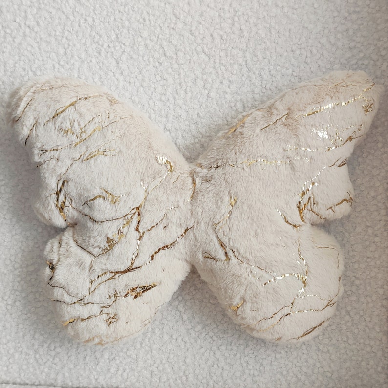 May include: A plush butterfly-shaped pillow in cream with gold metallic accents. The pillow has a soft, textured surface and a detailed design. The wings are spread out, and the gold accents create a striking contrast against the cream color.