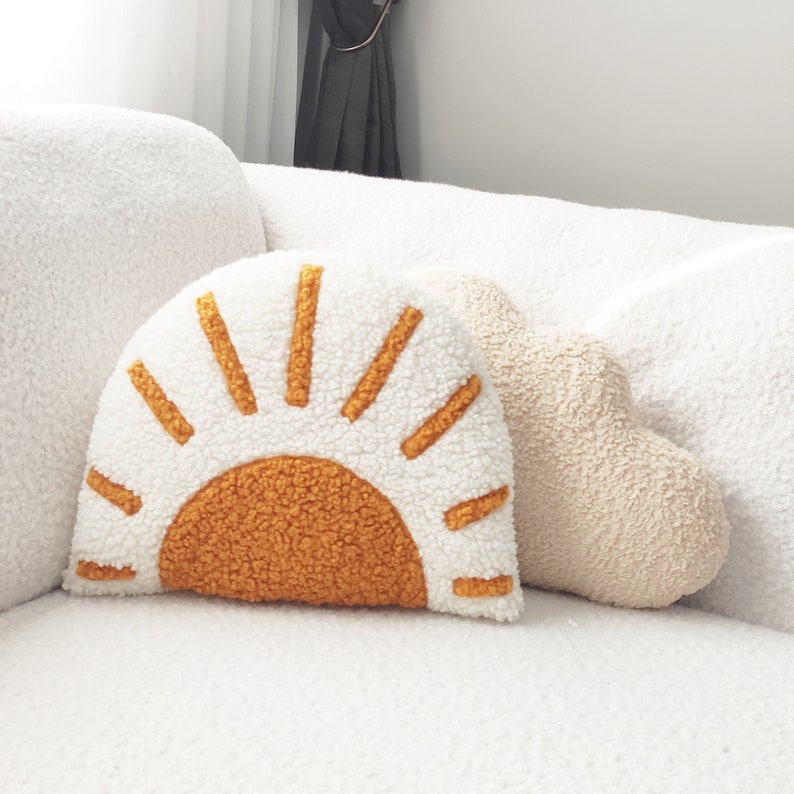 May include: Two decorative throw pillows. One is shaped like a sun with a white background and orange sun rays and center. The other is a beige cloud shape. Both are made of a soft, textured material.