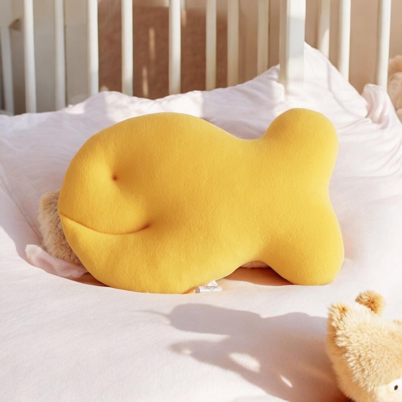 May include: A bright yellow, fish-shaped pillow rests on a white bed. The pillow has a soft, plush texture and a smiling face. The background includes a white crib and a small stuffed animal.