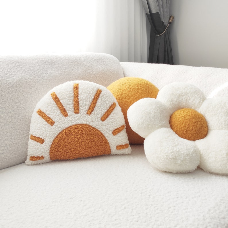 May include: A collection of decorative throw pillows in various shapes and colors. The pillows include a white and orange sunburst, a yellow sphere, and a white daisy with a yellow center. The pillows are made of a soft, textured material.