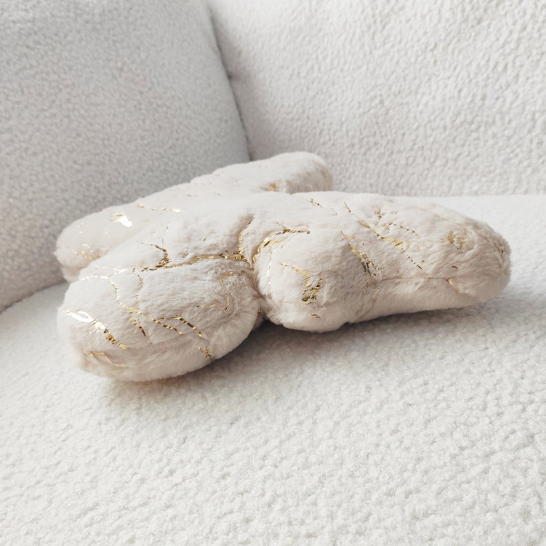May include: A plush, cream-colored decorative pillow with gold metallic accents. The pillow has a soft, textured surface and is shaped like a stylized cloud or abstract form. The gold accents create a subtle, elegant pattern across the surface.